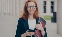 Portrait of confident beautiful red-haired female business consultant holding modern smartphone and laptop, looking at camera with smile, wearing spectacles, standing outdoors after working day